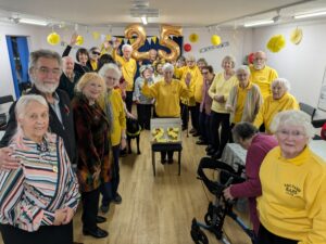 A group of about 20 people pose for a photo in a room decorated by balloons and bunting. Two of the balloons spell out 25. The woman in the middle of the group is wearing a yellow jumper with the words Saltash BAPS on it and in front of her is a cake in the shape of the numbers 25.