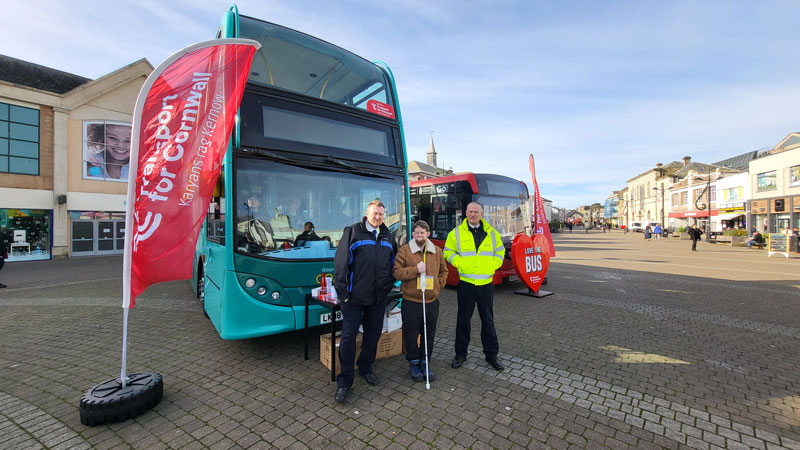 Three men pose for a photo outside, stood in front of two buses. One is a large double decker and the other is a standard bus. There is a large banner which reads Transport for Cornwall. One of the mean is wearing a lanyard which reads 'I am visually impaired' and the other two men have First Kernow and Transport for Cornwall branded clothes.