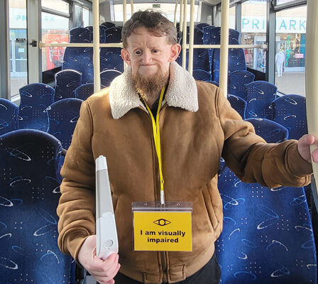 A man holding a white cane and wearing a lanyard which reads 'I am visually impaired' around his neck stands in the aisle of a bus. From the blog: How to catch a bus as a blind or partially sighted person