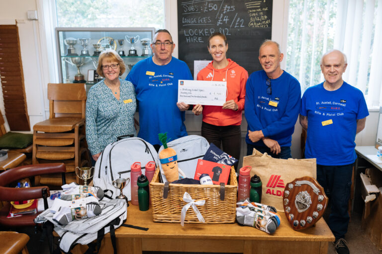 A group of people pose for a photo holding a giant cheque. The woman in the middle is wearing Team Great Britain merchandise whilst the three men and one woman standing either side of her are wearing iSightCornwall volunteer badges and Blind Using Guided Sights Club branded jumpers