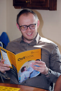 A man smiles as he reads a magazine which is titled Outlook and has the iSightCornwall Logo on it