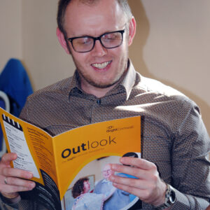 A man smiles as he reads a magazine which is titled Outlook and has the iSightCornwall Logo on it