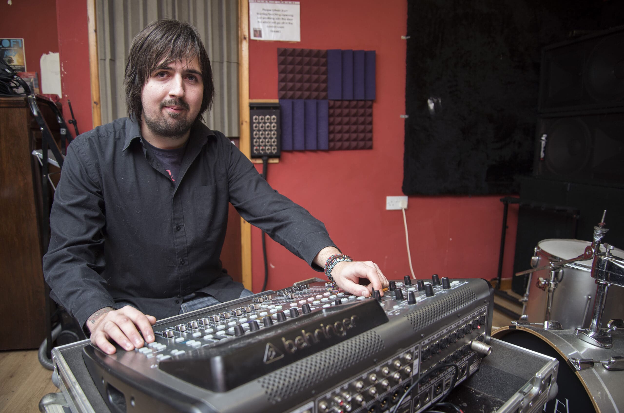 A man with dark hair and a beard sits in front of a mixing desk with his hands on some of the dials.