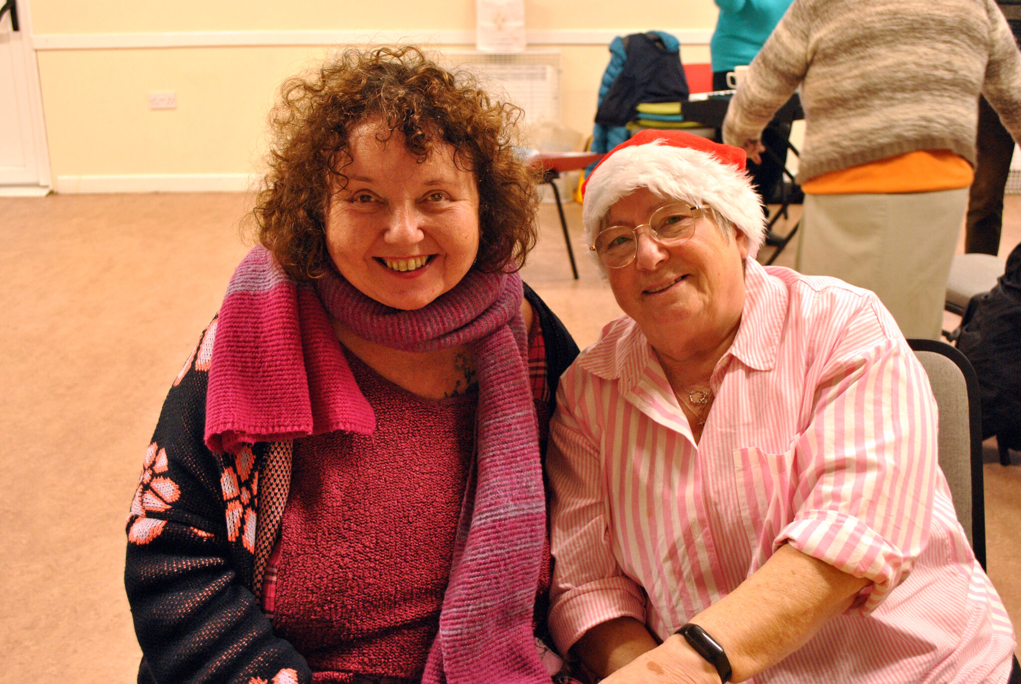 Two women smile for the camera. One is wearing a Santa hat and the other has a colourful scarf