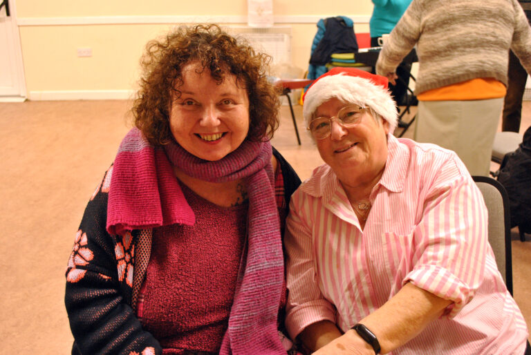 Two women smile for the camera. One is wearing a Santa hat and the other has a colourful scarf