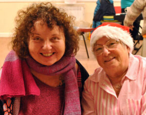 Two women smile for the camera. One is wearing a Santa hat and the other has a colourful scarf