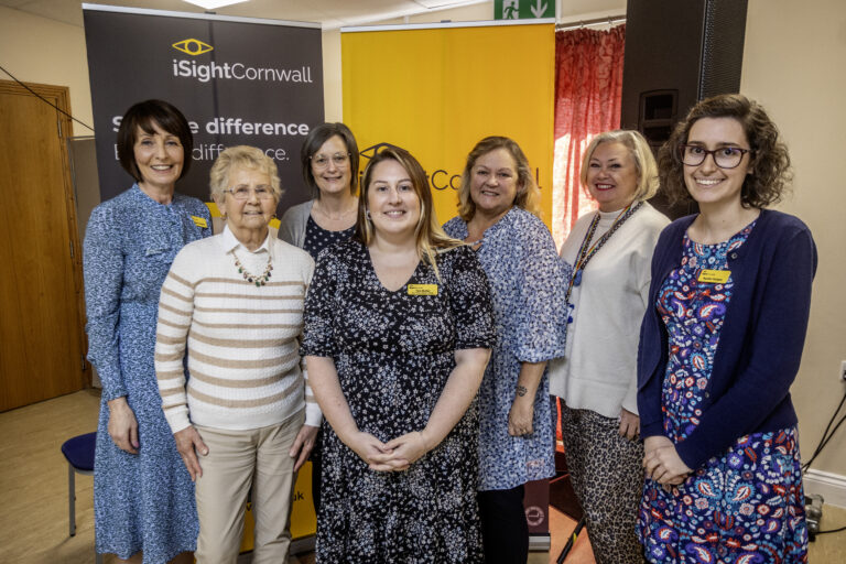 A group of 7 women smile for the camera, whilst posing in front of an iSightCornwall banner.