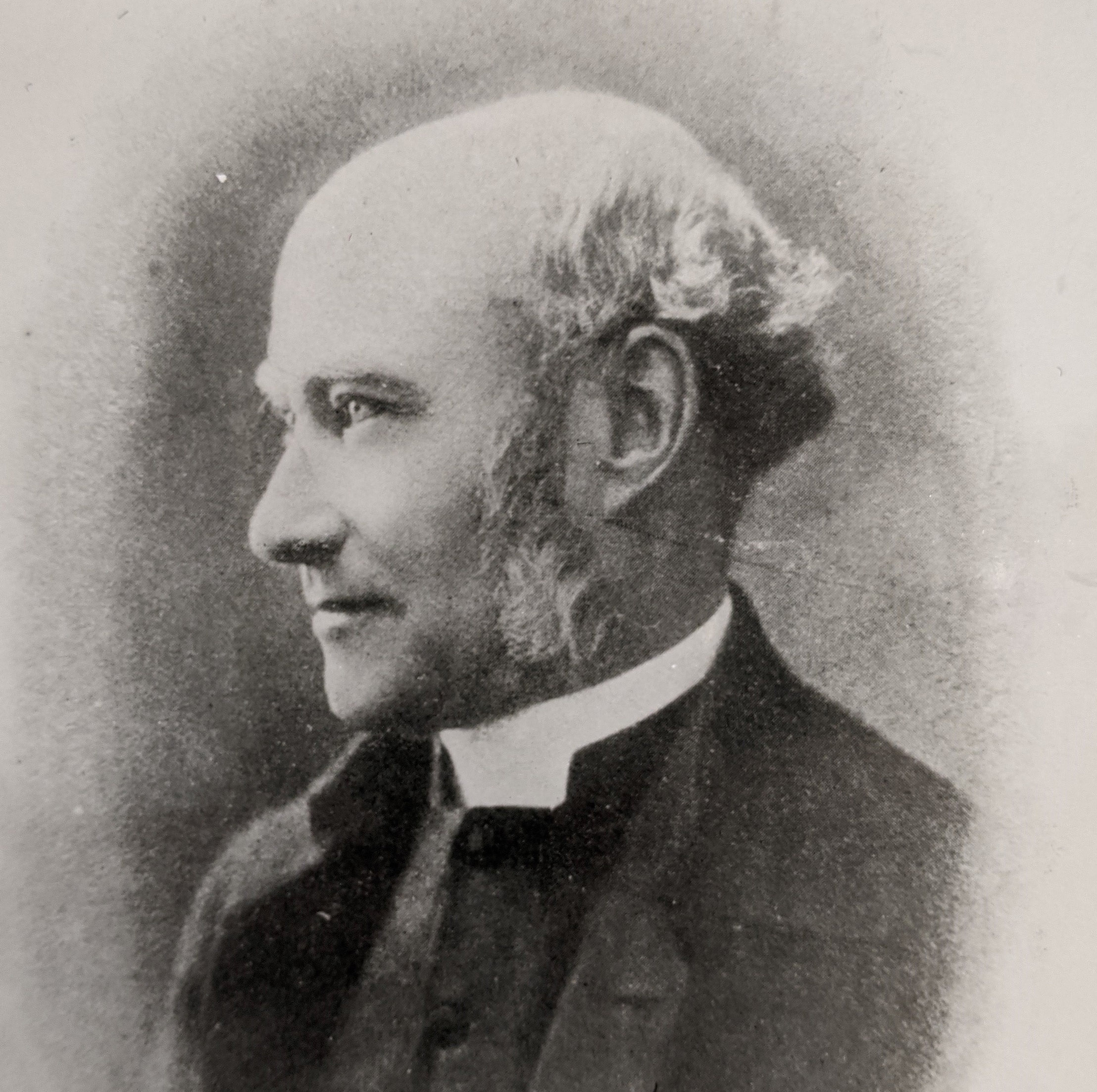 A black and white image of a man with a receding hairline and large sideburns wearing a clerical collar. He is in side profile looking to the left.