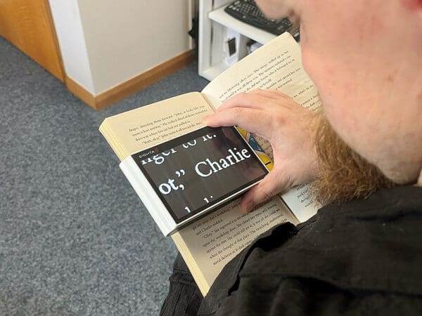 A handheld device with a screen is held over a book, the text from the book is shown on the screen in a much larger font