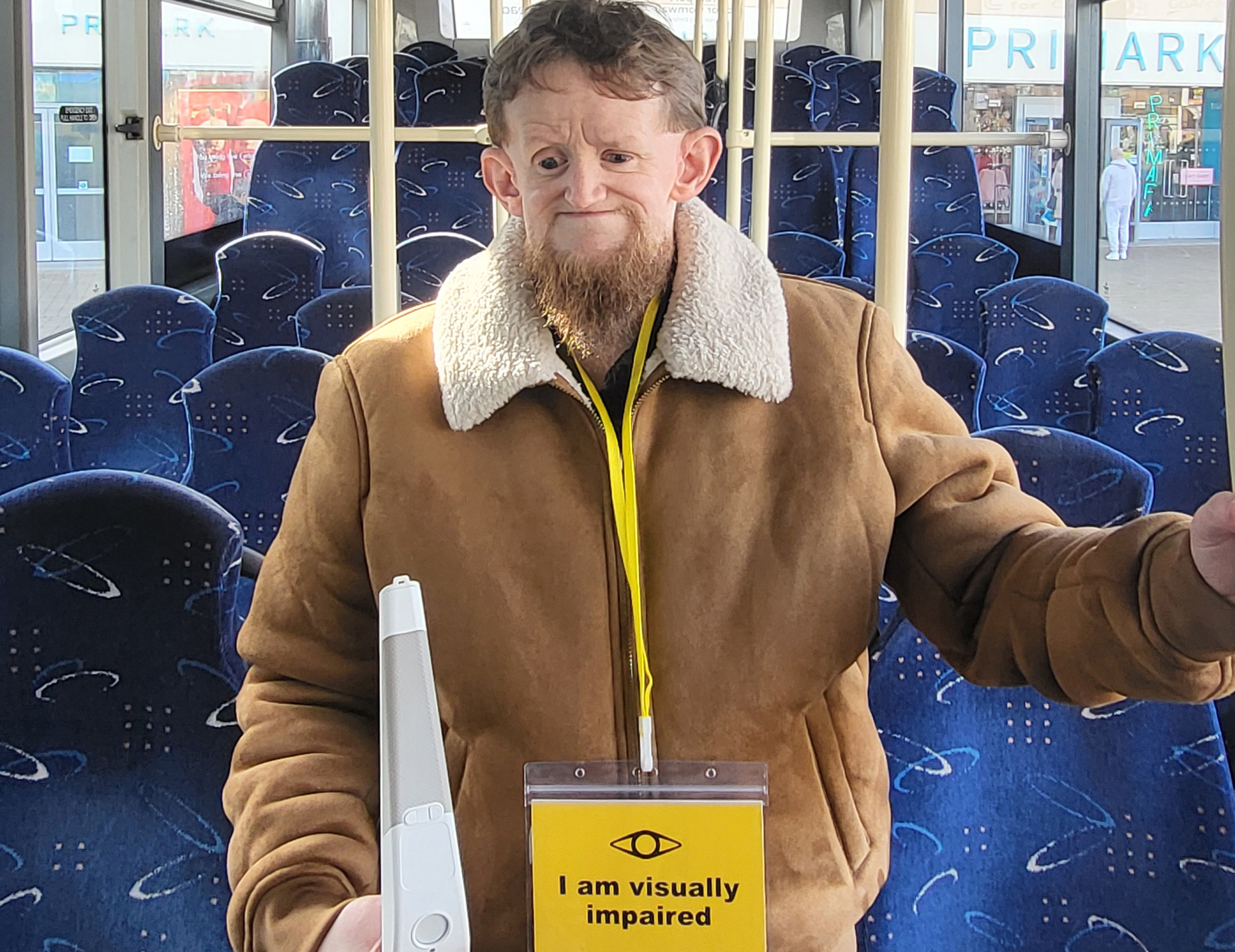 A man holding a white cane and wearing a lanyard which reads 'I am visually impaired' around his neck stands in the aisle of a bus.