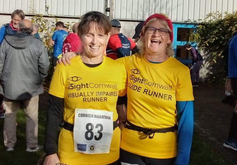 Two women in yellow iSightCornwall t-shirts stand next to each other smiling and laughing. One woman's t-shirt says guide runner and she has her arm around the other woman whose t-shirt says visually impaired runner.