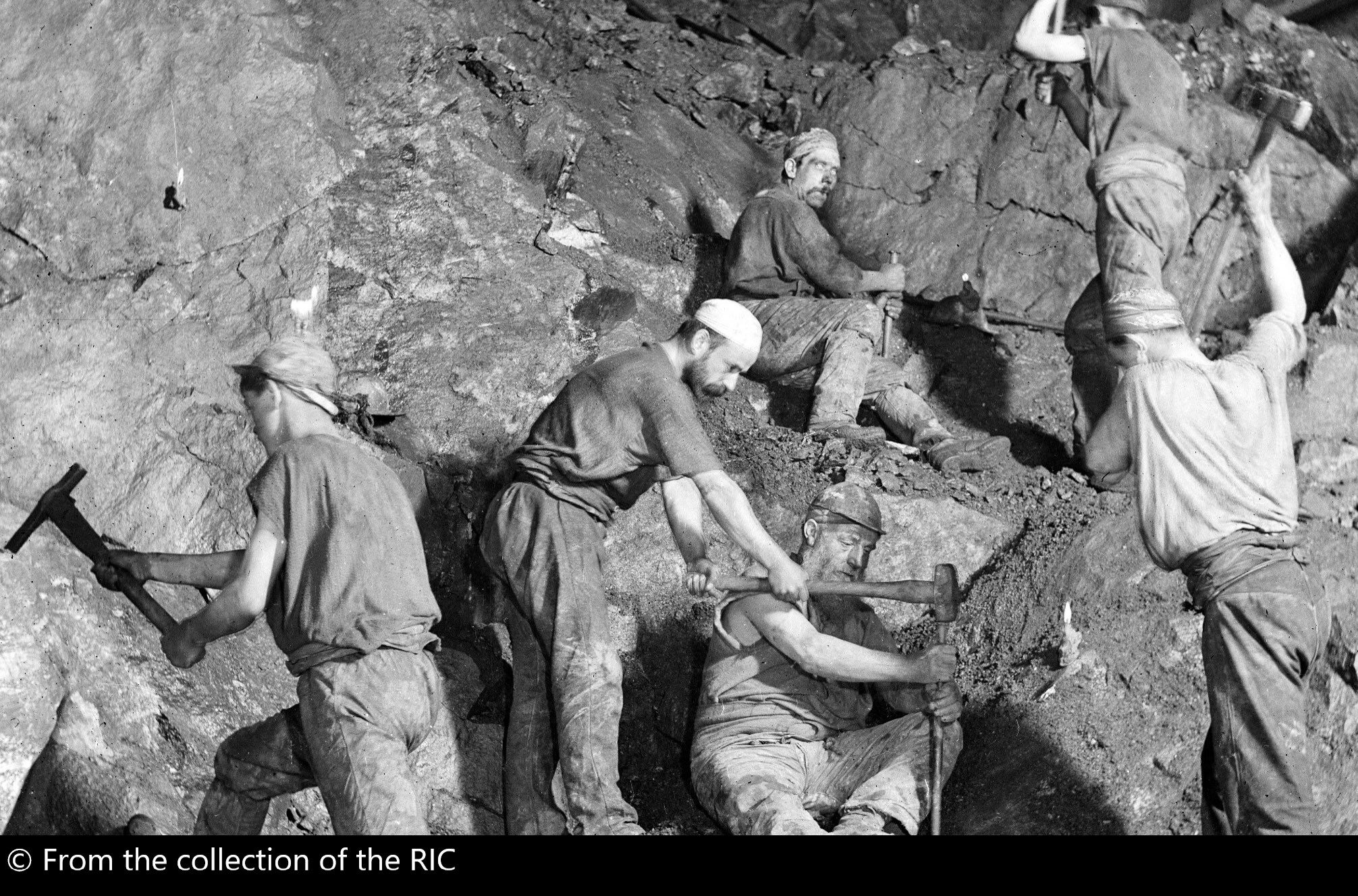 Black and white photo of miners working underground wearing helmets and holding tools.