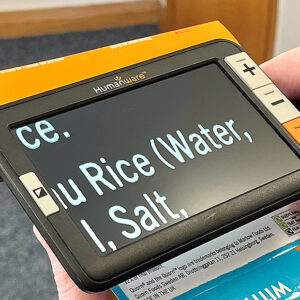 an image showing an electronic magnifier with the Humanware logo on it. It has a screen and four buttons. The buttons have a plus, a minus and an image of a camera. The screen is showing an enlarged version of a recipe. The device shown is the Humanware Explore 5