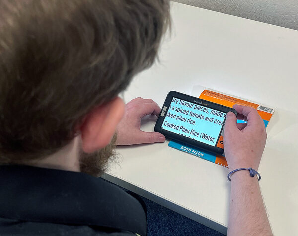 an image showing an electronic magnifier with the Humanware logo on it. It has a screen and four buttons. The buttons have a plus, a minus and an image of a camera. It is being held by a man who's back of head and hands are visible as he used the magnifier to increase the size of the text of a document he is reading. Device shown is the Humanware Explore 5