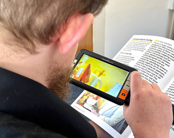 an image showing an electronic magnifier with the Humanware logo on it. It has a screen and four buttons. The buttons have a plus, a minus and an image of a camera. It is being held by a man who's back of head is visible as he used the magnifier to increase the size of the text of a magazine he is reading. Device shown is the Humanware Explore 5