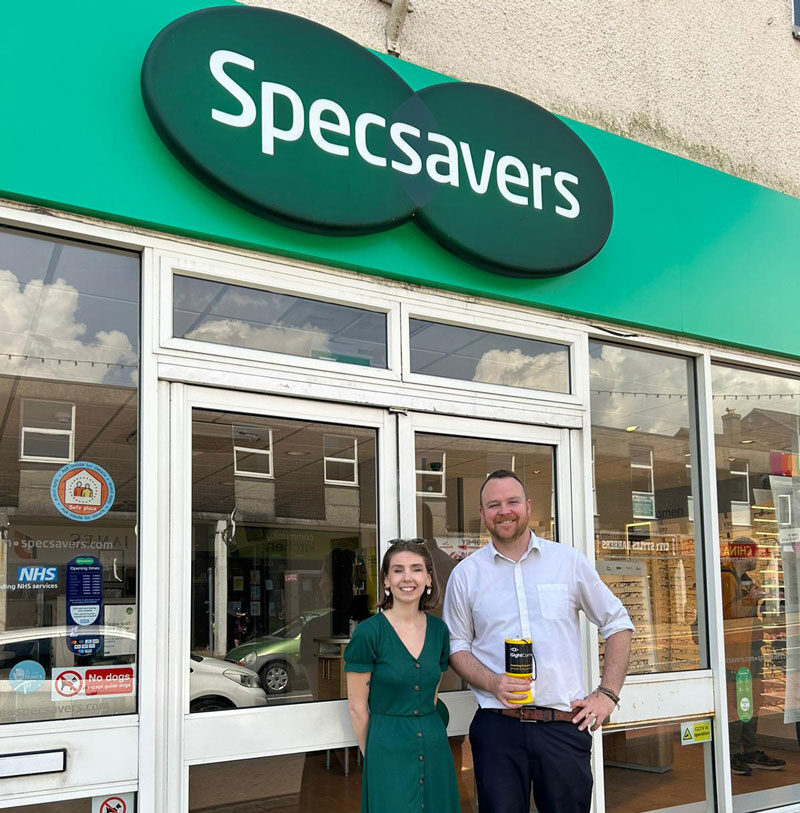 A man and woman stand outside a Specsavers shop whilst holding an iSightCornwall collection tin