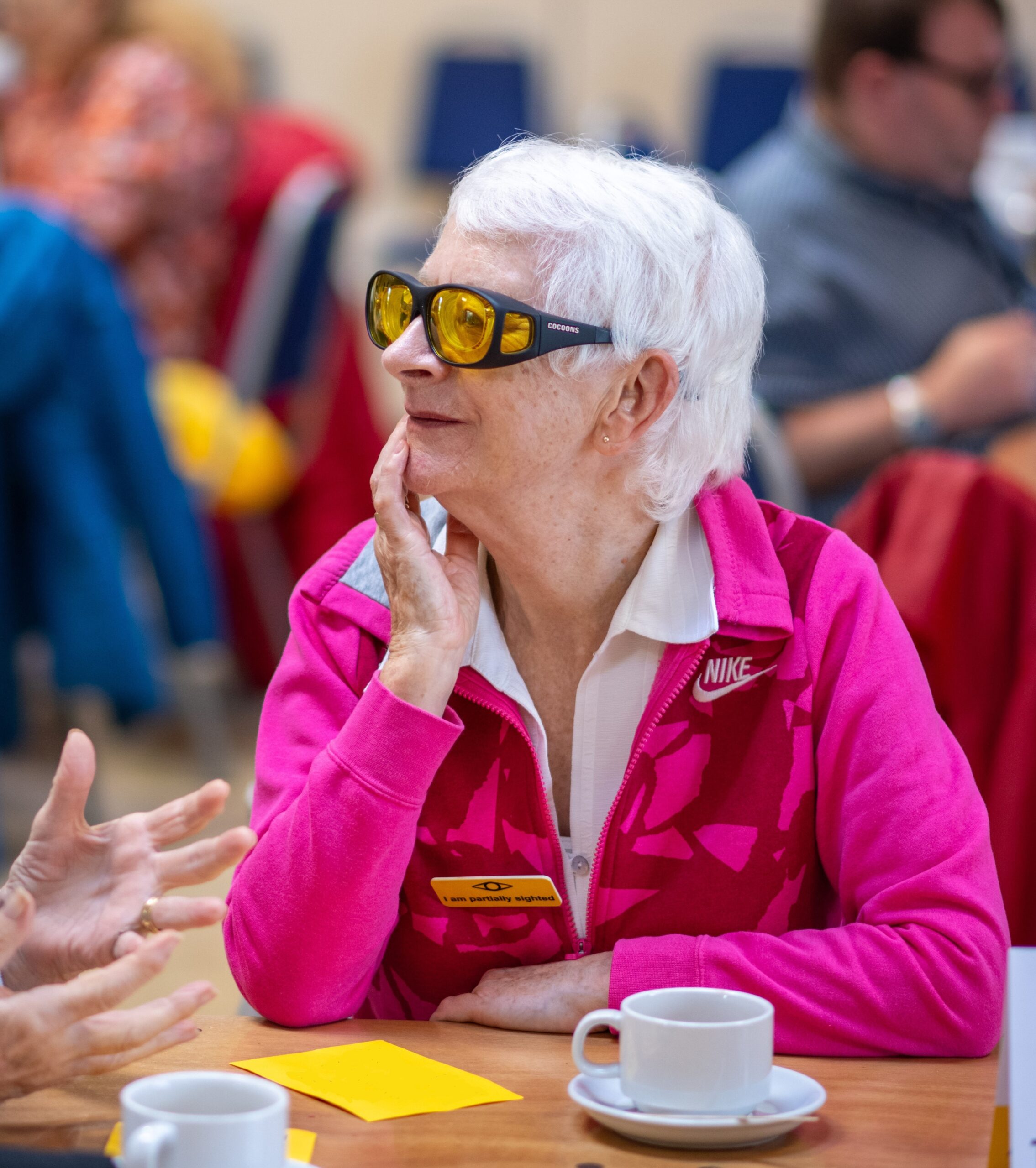 a woman wearing filter glasses and a badge which reads I am Partially Sighted sitting at a table with a cup and saucer in front of her. She is listening intently to someone off camera.