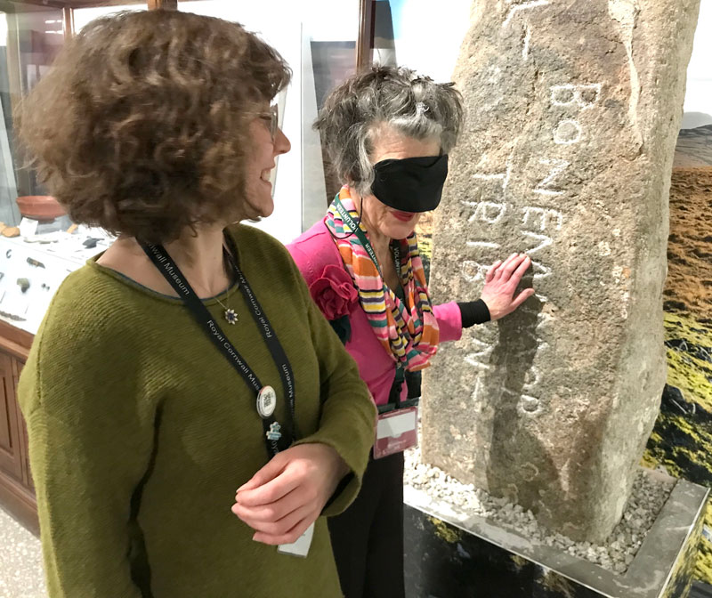 A woman wearing a lanyard which reads Royal Cornwall Museum smiles as she guides another woman wearing a blindfold around what looks like an old artifact