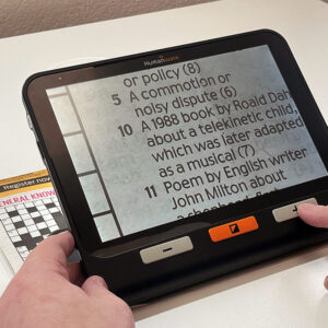 A large rectangular electronic device with a screen and three simple buttons. The screen is showing an enlarged image of a newspaper crossword which is placed underneath the device. The device is the Humanware Explore 8 Electronic Magnifier