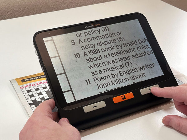 A large rectangular electronic device with a screen and three simple buttons. The screen is showing an enlarged image of a newspaper crossword which is placed underneath the device. The device is the Humanware Explore 8 Electronic Magnifier