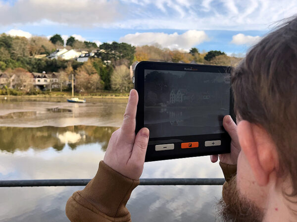 A large rectangular electronic device with a screen and three simple buttons. It is being held by a man who is pointing it at a nice harbour scene outside of the window and the device is enlarging the view on the screen. The device is the Humanware Explore 8 Electronic Magnifier