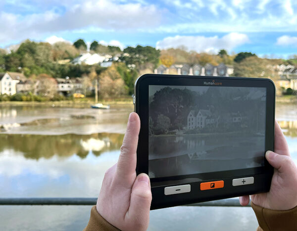 A large rectangular electronic device with a screen and three simple buttons. It is being held by a man who is pointing it at a nice harbour scene outside of the window and the device is enlarging the view on the screen. The device is the Humanware Explore 8 Electronic Magnifier