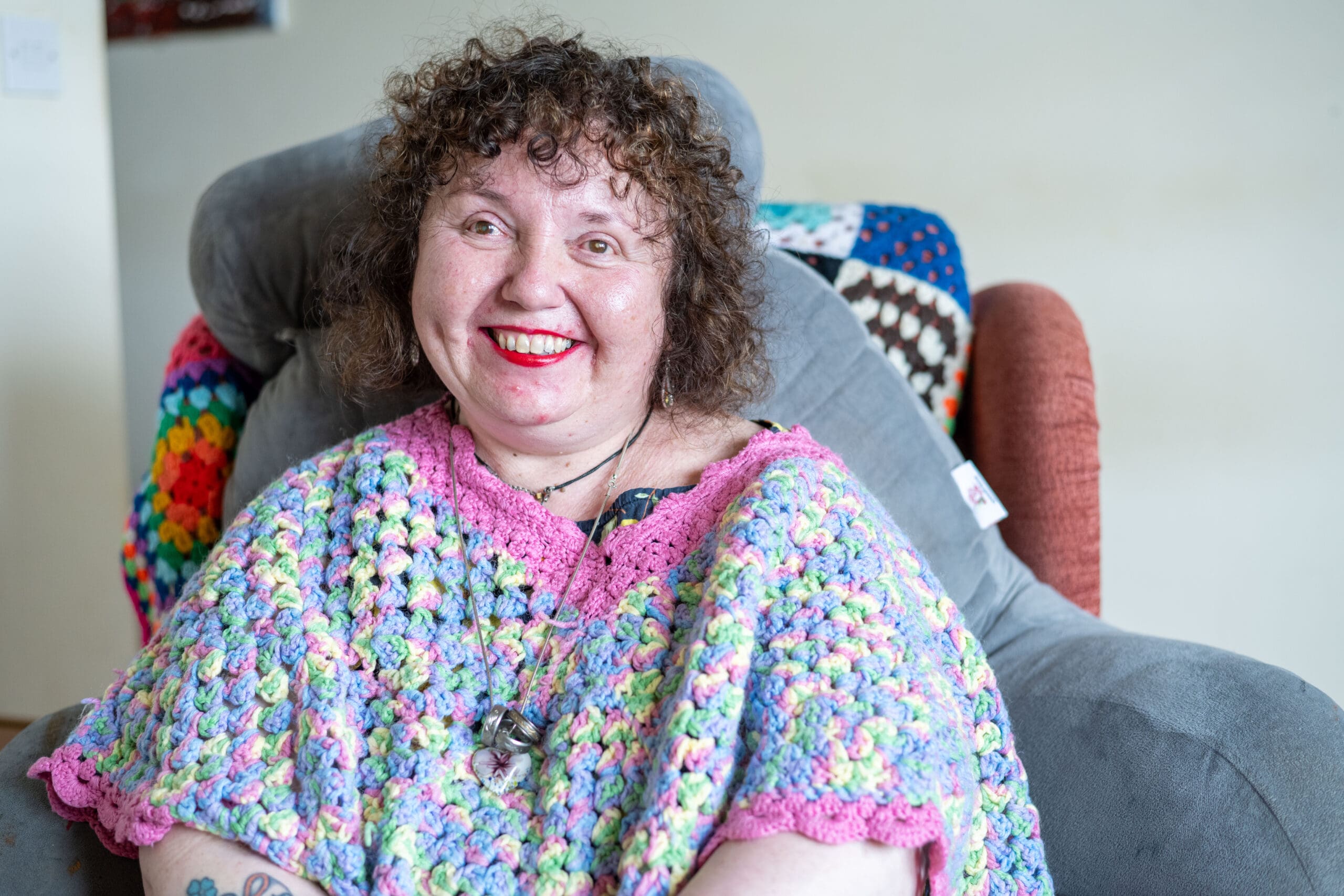 A woman wearing a brightly coloured shawl smiles at the camera. She is sitting in an armchair in what appears tp be a living room.