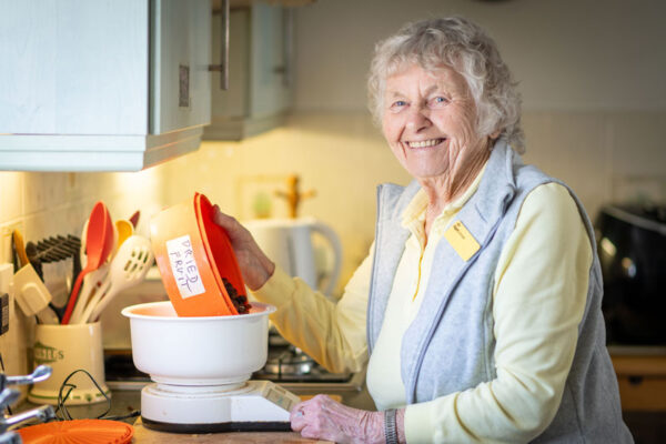 A woman wearing an iSightCornwall badges smiles as she empties a bowl of dried fruit into a kitchen scale