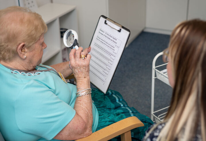 Lady in a turquoise top sitting in a chair and holding a magnifier to read a chart.