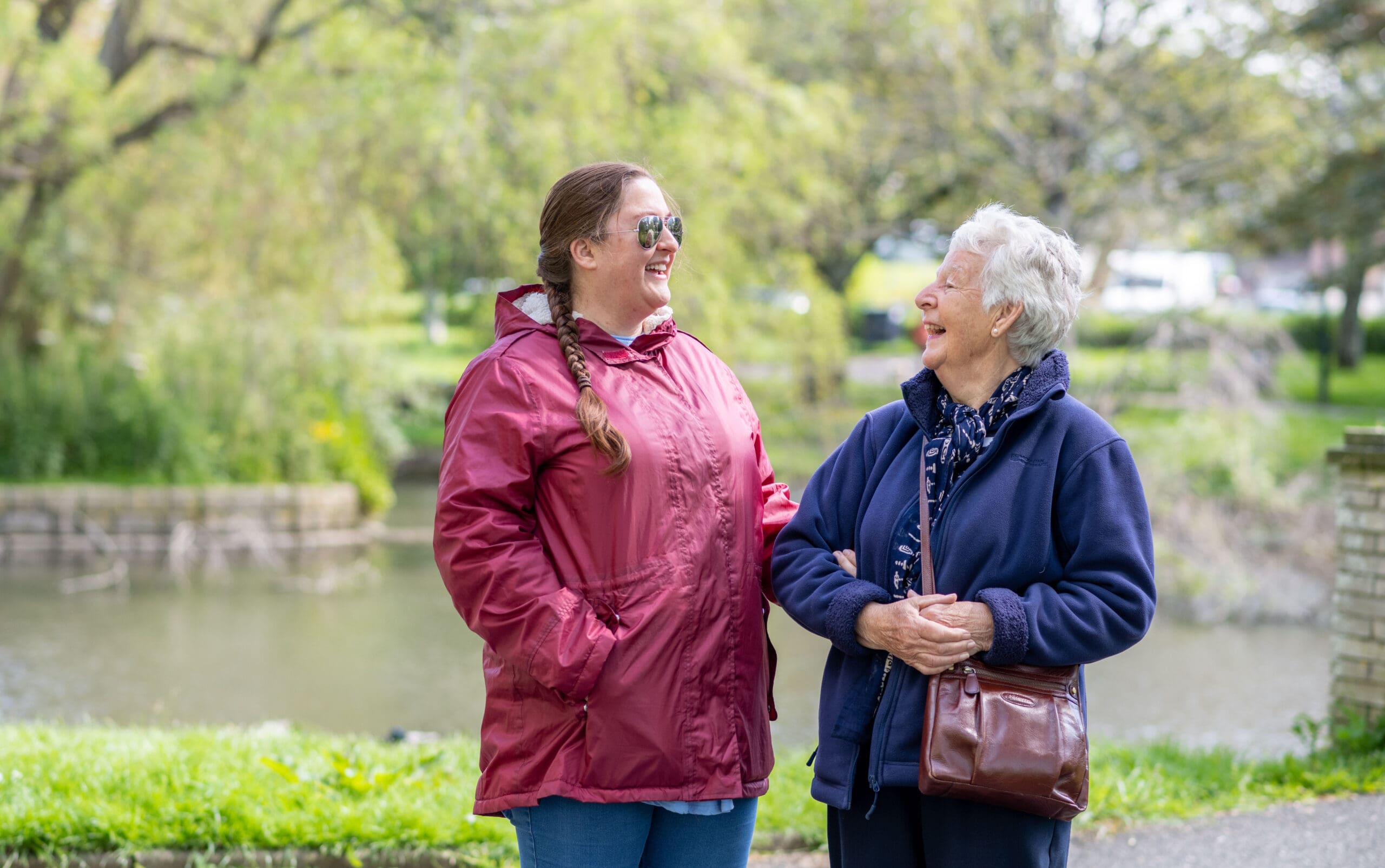 Two ladies stand arm in arm in front of a lake. They are looking towards each other and smiling.