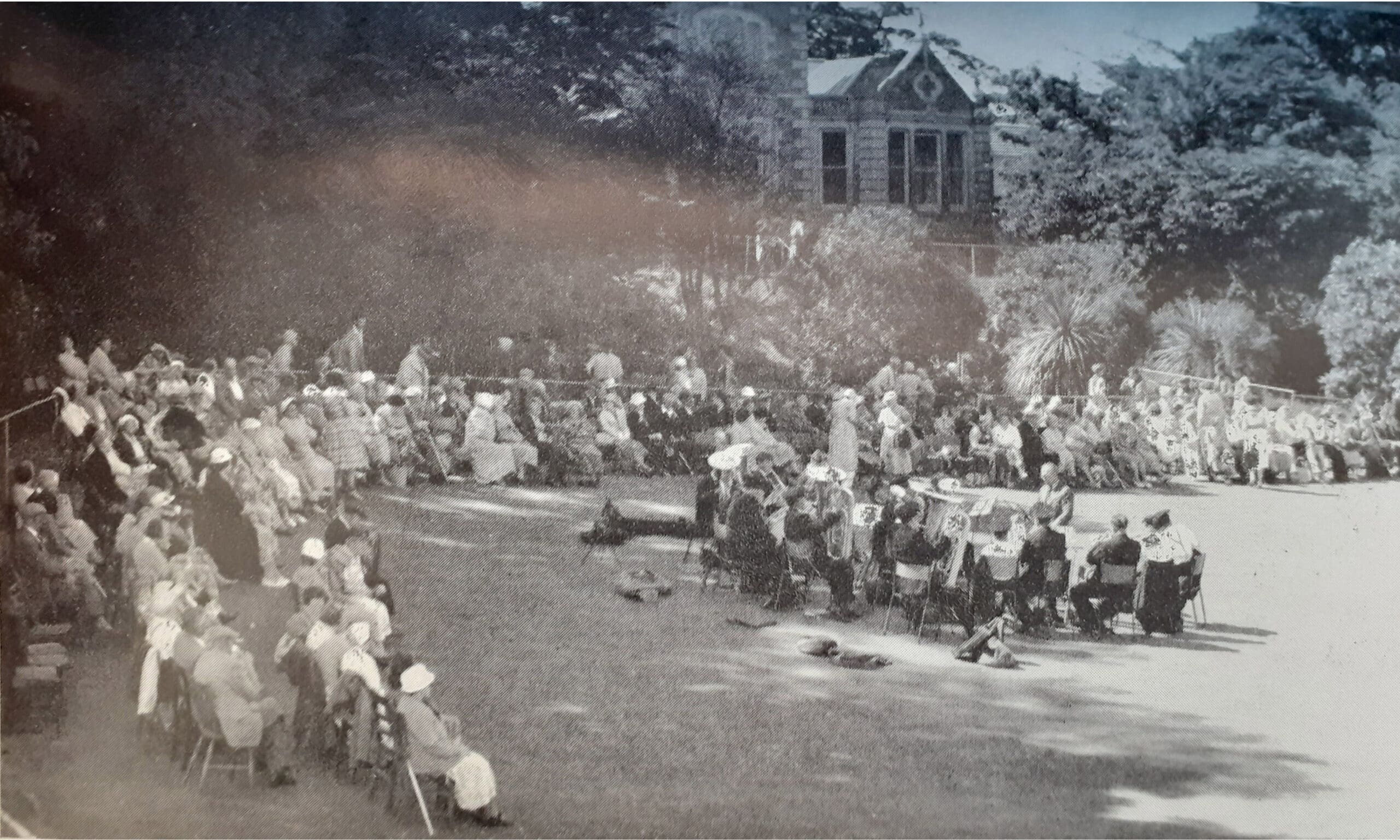 A grainy black and white image showing approximately 100 people sat on chairs on a lawn outside a large house. In the foreground is an orchestra and conductor.