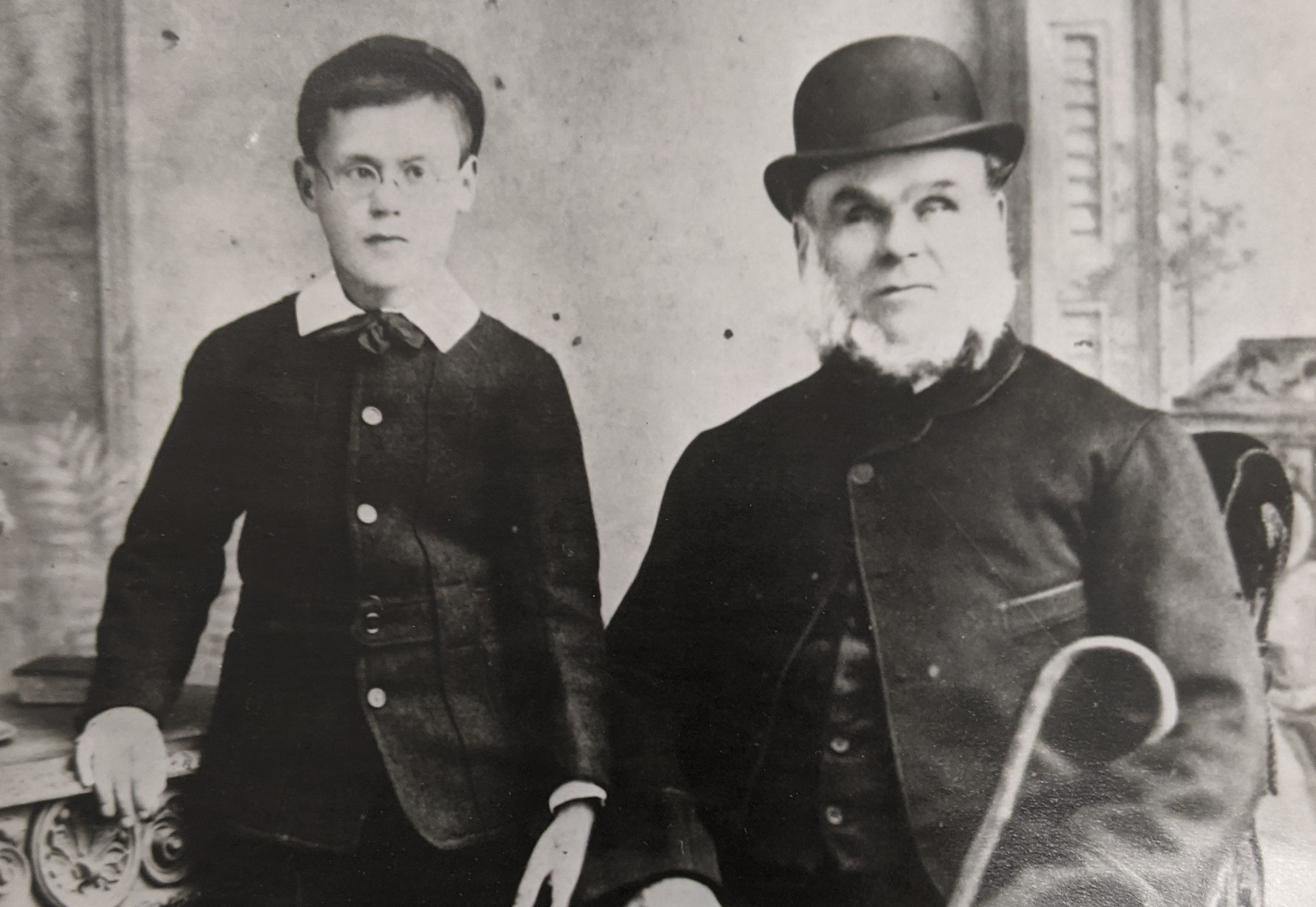 A black and white image of a young boy wearing glasses stands next to an older gentleman wearing a bowler hat.