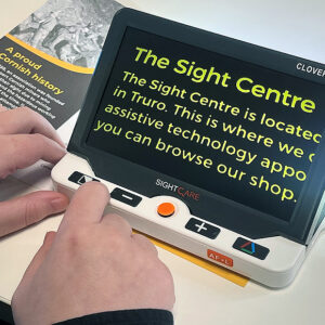 A large rectangular electronic device with a screen and five buttons. The device has a built in stand and is sitting on top of a leaflet. The text from the leaflet is shown on the screen of the device in an enlarged font. The device is the Optima Clover 7S