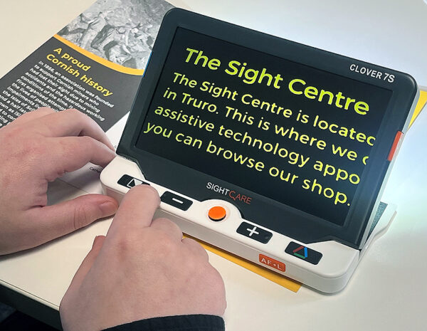 A large rectangular electronic device with a screen and five buttons. The device has a built in stand and is sitting on top of a leaflet. The text from the leaflet is shown on the screen of the device in an enlarged font. The device is the Optima Clover 7S