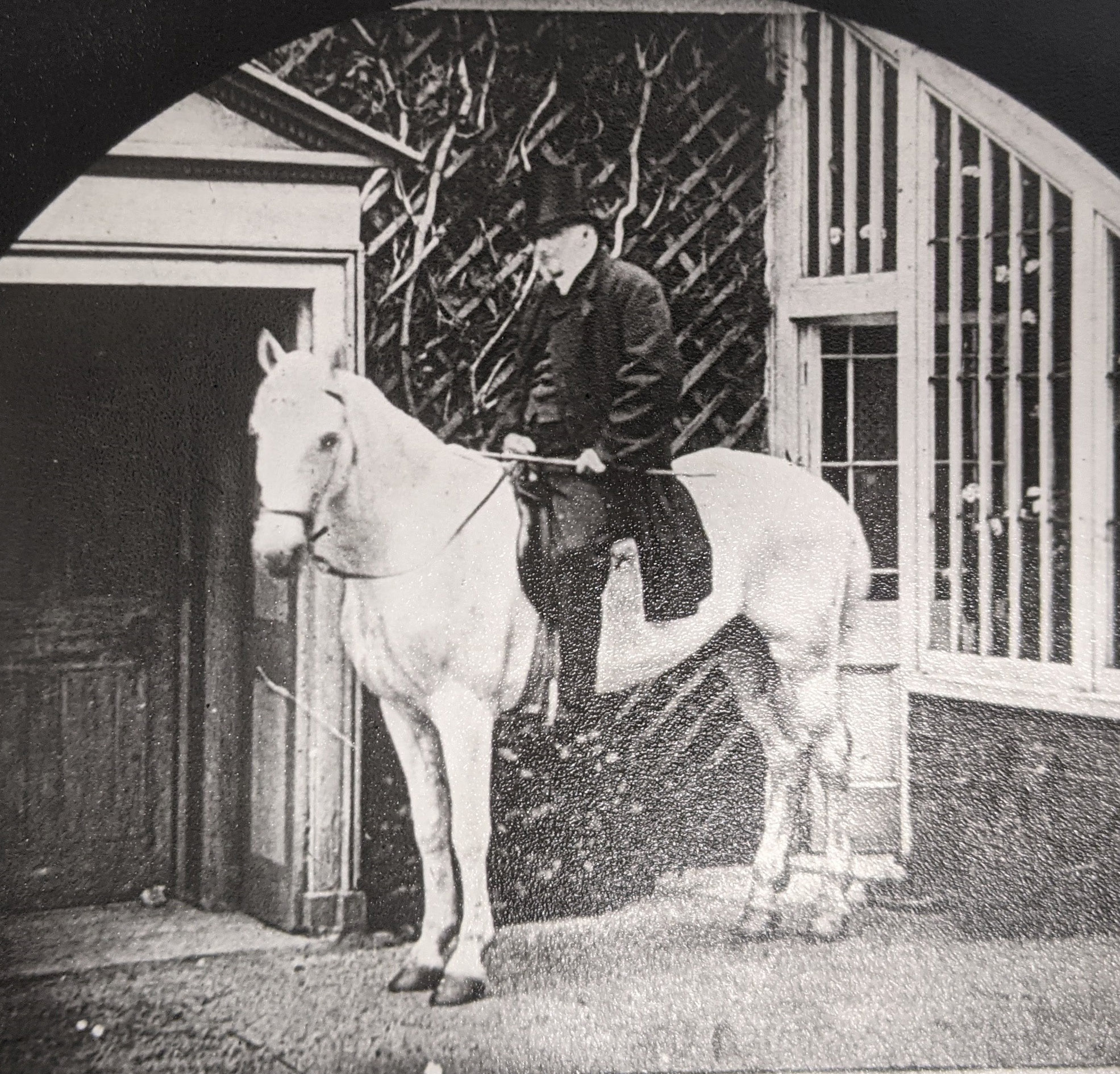 A grainy black and white photograph of a man in top hat and tails sitting on a white horse outside a grand looking building.