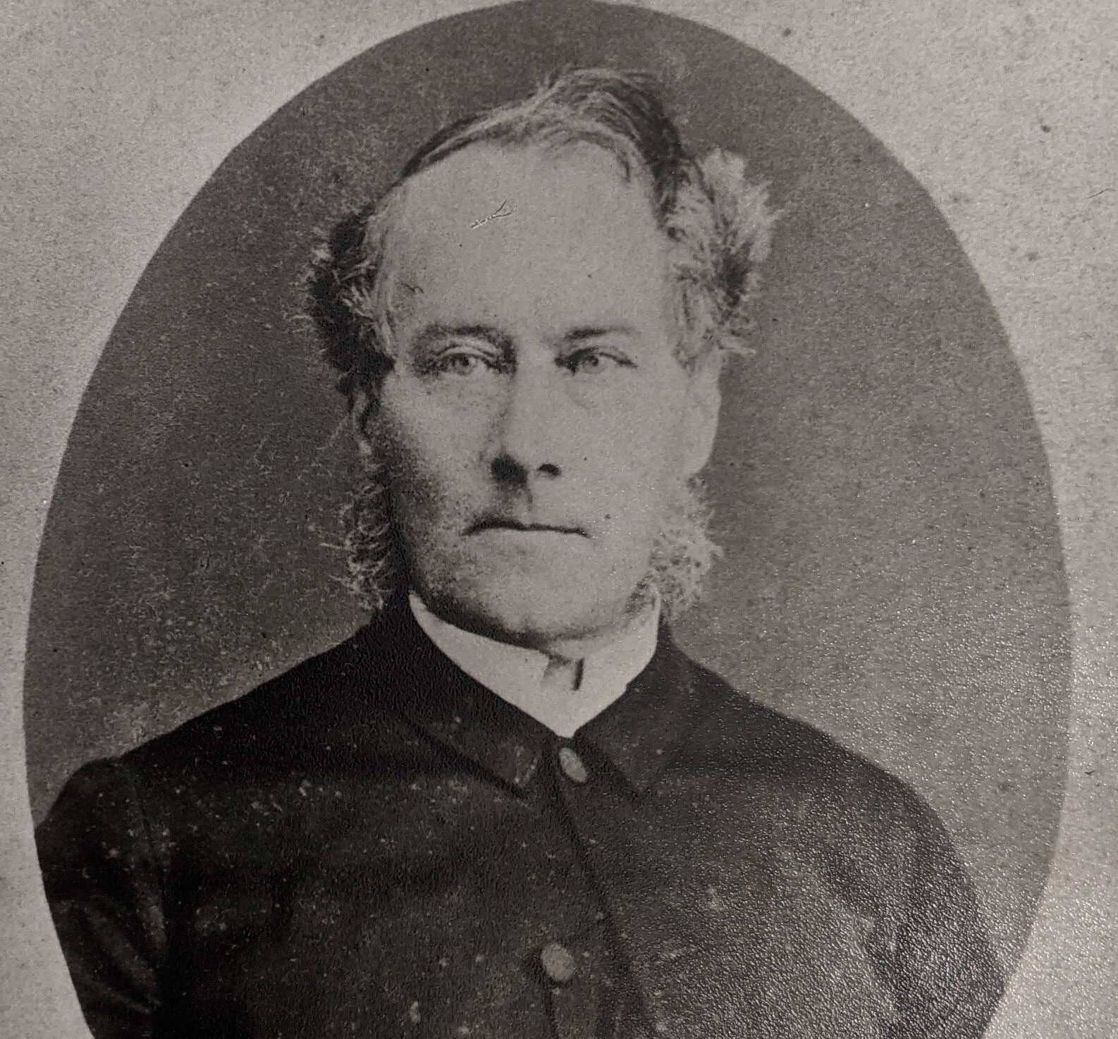 A black and white image of a man with large side burns wearing a buttoned up black jacket.