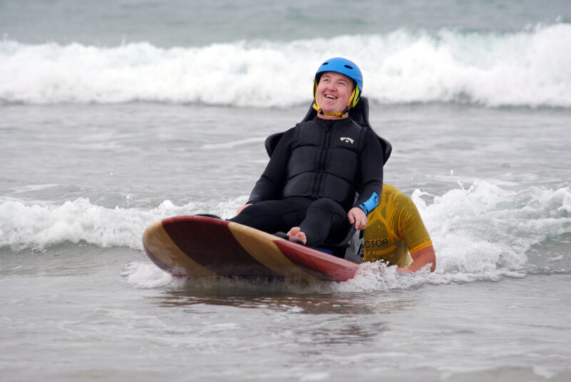 A young man sits on a surf board in the water. He's wearing a helmet and smiling as he rides a wave, helped by a man holding the board behind him.