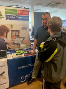 Dom from iSightCornwall speaks to a representative at the Optelec table. Behind them on the table is a range of electronic magnifiers and other devices.