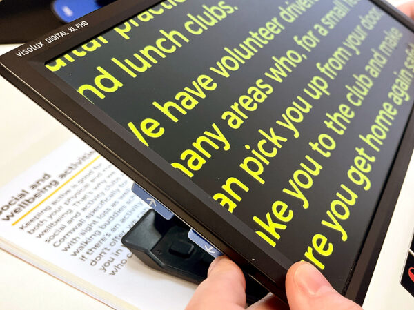 The side view of a large rectangular device with a screen being held above a book. The text in the book is being shown on the screen of the device in a larger font and different colour