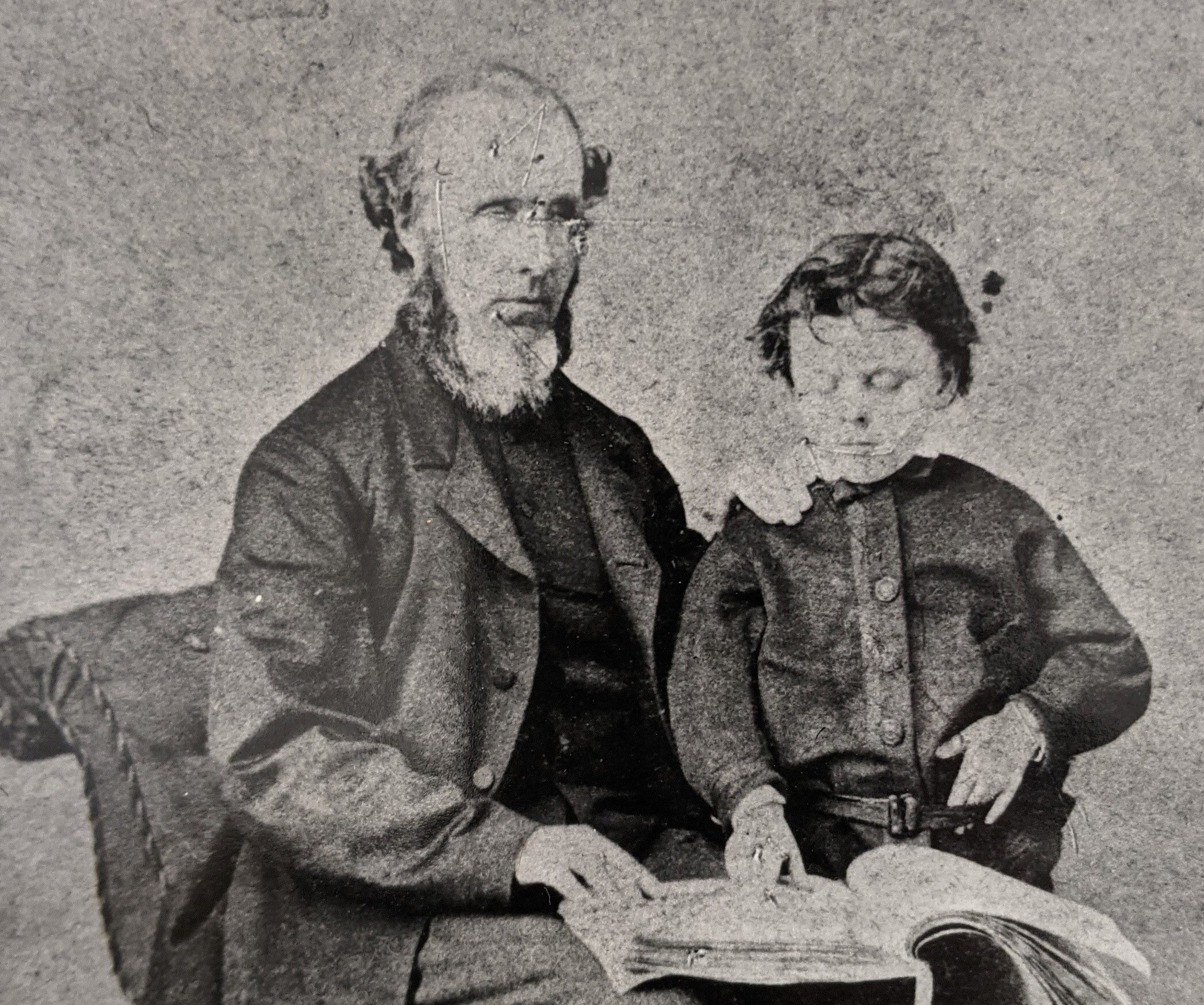 A grainy black and white photo of a man sitting down with a book on his knee. Standing next to the man is a young boy with his hand on the book.