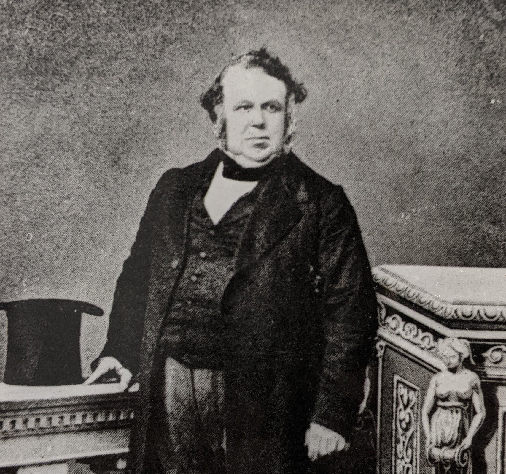 A black and white photo of a portly gentleman with impressive side burns. He is wearing coat tails and is standing next to a plinth which is holding his top hat.