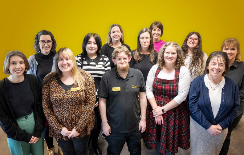 The 12 members of the iSightCornwall team standing in front of a yellow background.