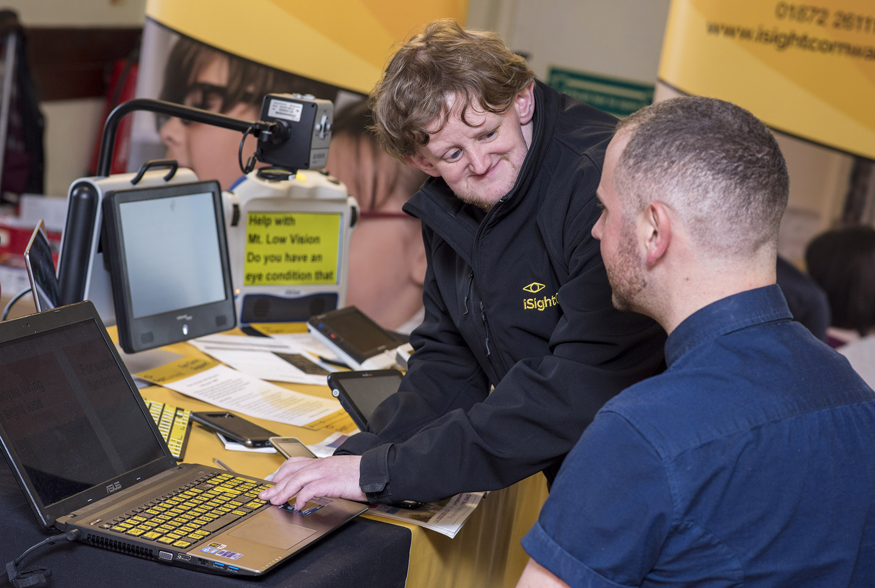 Two men are interacting with a computer that has a magnifier on it enlarging the text. One of the men is wearing an iSightCornwall name badge and seems to be demonstrating something to the other man